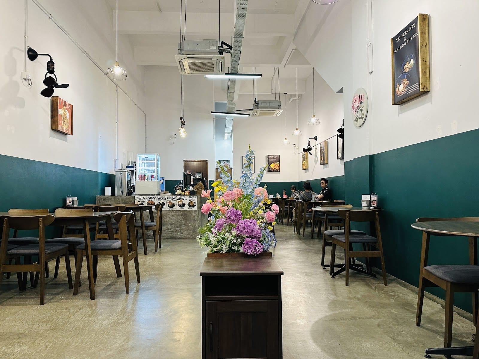 Riceater's dining room: white walls with a deep green lower half, wooden tables and chairs, hanging lamps and a large flower arrangement down the middle
