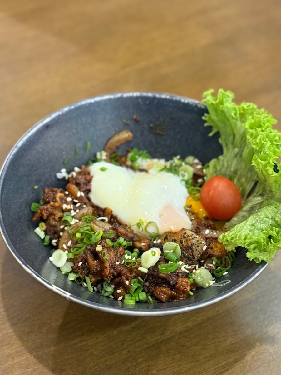 A bowl of pork topped with a soft egg, spring onion, sesame seeds and a cherry tomato